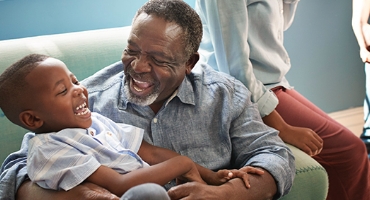 Black child sitting on bearded grandfather's lap, both are laughing, someone sits nearby; represents Highmark Medicare Plans
