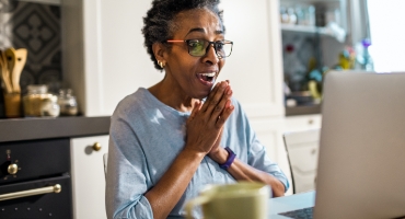 Older black woman sits at kitchen table at laptop, hands clasped, looking delighted reading about Highmark Medicare Plans 