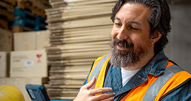 A man wearing a hard hat and safety vest smiling while looking at a tablet in what appears to be a warehouse setting.