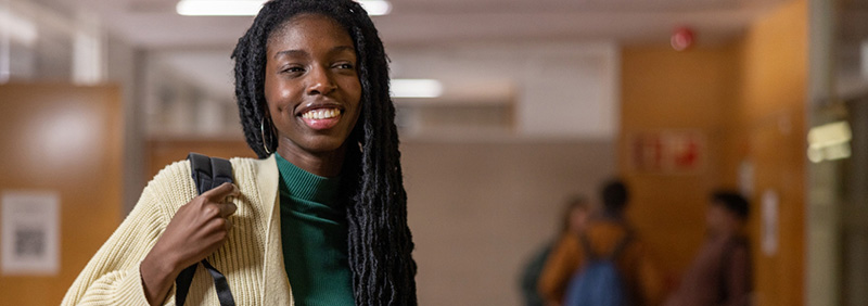 young female smiling holding a backpack on one shoulder standing in a hallway of a school