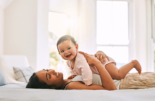 a smiling woman lying on a bed with a smiling baby laying on her chest