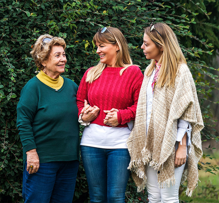 Older woman with two younger women, possibly caregivers or family, standing outside, smiling and holding hands.
