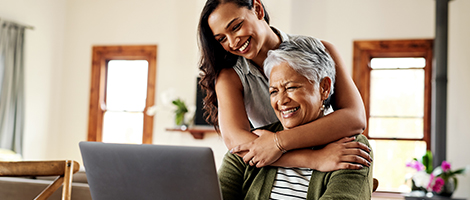 a woman standing behind her seated mother with her arms around her, both are smiling and looking at a laptop computer