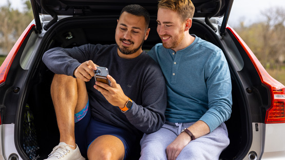 two men sitting in the back hatch of a car, smiling and looking at a cell phone together