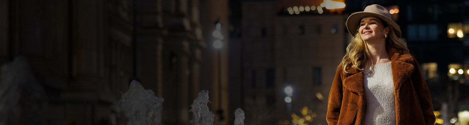 A person who looks to be in their 20s, wearing a brown furry coat and a hat, is smiling and looking up at something in the city of Philadelphia with lights on the buildings and fountain.