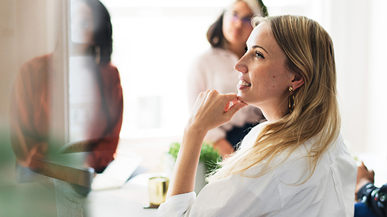 A women with blonde hair sits at a table, looking thoughtful during a meeting with other people in an office.