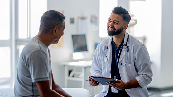 A smiling medical professional wearing a white coat and blue scrubs sits and talks with a patient while holding a tablet.