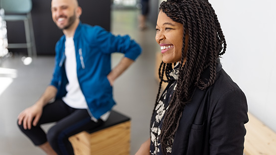 A person with long braids smiles while sitting, and another person sits behind them, smiling too.