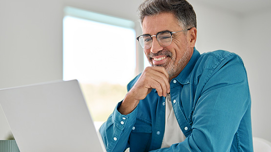 A smiling man with glasses and a blue shirt looks at a laptop screen.
