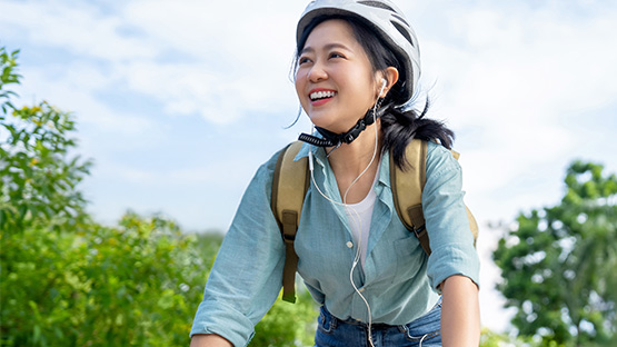 A young person with a happy expression is wearing a bike helmet and backpack while outdoors with green trees and a blue sky in the background.