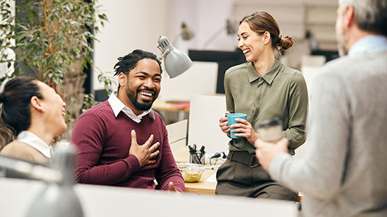 Four adults are laughing together in an office setting.