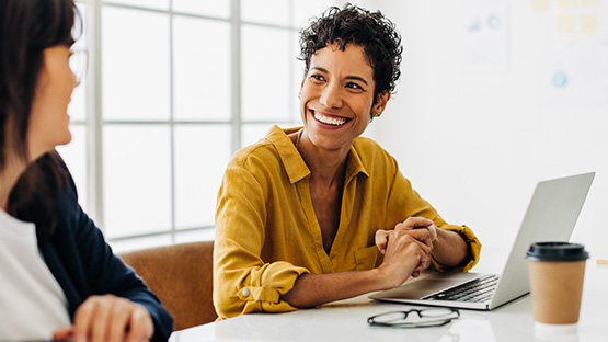 A smiling woman with short, curly hair is looking to her left and appears to be in conversation with another person. She is wearing a yellow collared shirt and is seated at a white table with a laptop, a coffee cup, and a pair of glasses beside her.