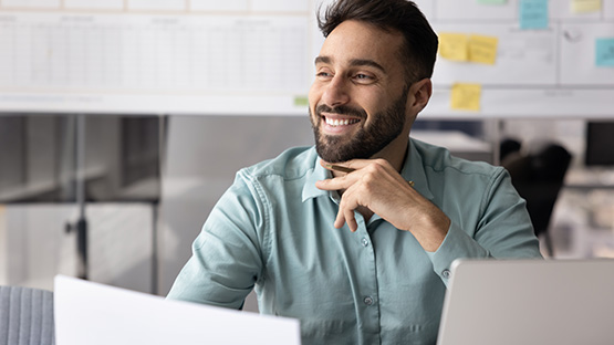 A happy man with a beard, in his 30s, smiles and looks to the right while sitting at a desk with a laptop.