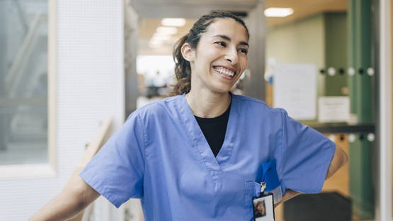A smiling medical professional in scrubs stands in a hallway and looks happy to be at work.