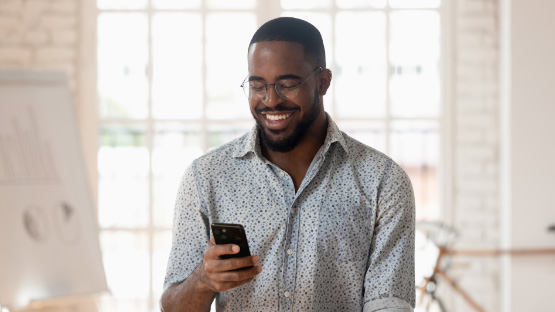 A smiling man who looks to be in his 30s wears glasses and a patterned shirt as he looks at his phone in a bright, modern office space.
