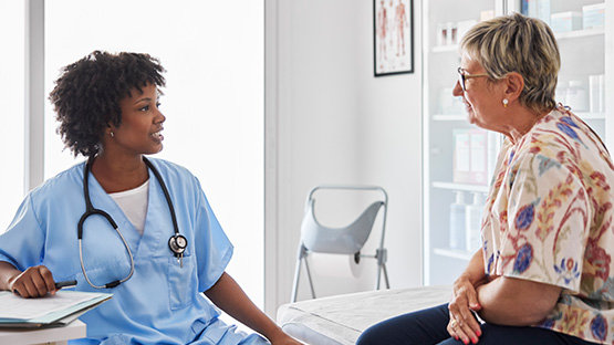 A medical professional in light blue scrubs and a stethoscope talks with a senior woman in a doctor's office.