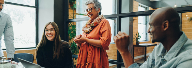 A person with short gray hair, wearing an orange dress and patterned scarf, laughs with hands clasped as younger coworkers smile nearby in a modern office.