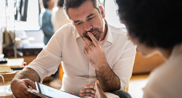 A person in a white collared shirt looks thoughtfully at a tablet held by another person while discussing something in a bright office.