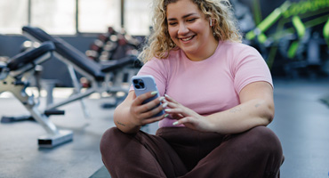 A young adult with curly blonde hair sits on the floor of a gym, smiling and looking at a phone.