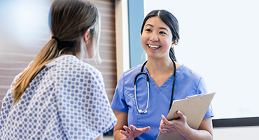 A smiling medical professional in a white lab coat and a medical professional in blue scrubs talk to a patient in a medical gown, offering comfort and support.