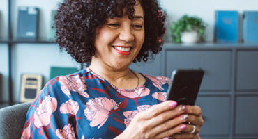 A smiling woman with curly hair looks at their phone in a room with a bookshelf
