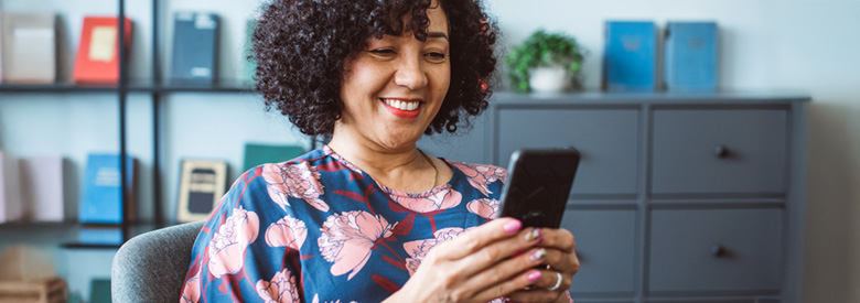 A smiling woman with curly hair looks at their phone in a room with a bookshelf