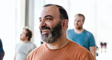 A bearded man in an orange t-shirt smiles while looking to the right, with other blurred figures in the background. 