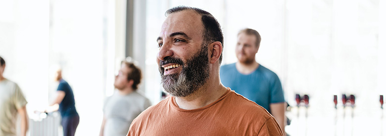 A bearded man in an orange t-shirt smiles while looking to the right, with other blurred figures in the background. 