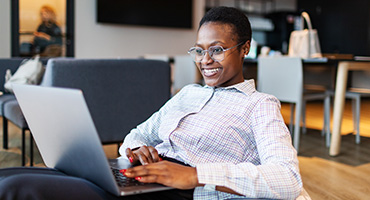 A smiling Black woman with short hair and glasses uses a laptop in a relaxed office setting, wearing a plaid shirt.