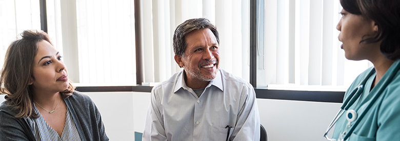 A smiling man and a woman in a grey cardigan listen attentively to a healthcare professional in turquoise scrubs.