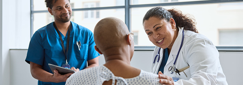 a medical professional in a white coat with a stethoscope and another medical professional in blue scrubs smile kindly at an adult patient in a hospital gown