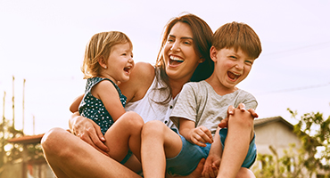 A mother and her two young children, a girl and a boy, are laughing joyfully outdoors, embodying family mental health and happiness.