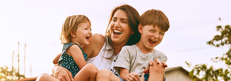 A mother and her two young children, a girl and a boy, are laughing joyfully outdoors, embodying family mental health and happiness.