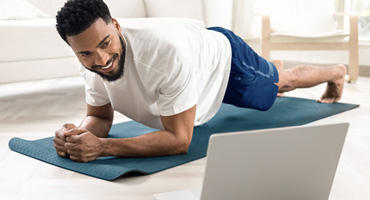 A person in their 20s or 30s is in a plank position on a yoga mat, smiling while watching a computer in a living room.
