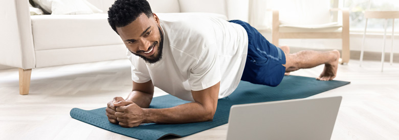 A person in their 20s or 30s is in a plank position on a yoga mat, smiling while watching a computer in a living room.