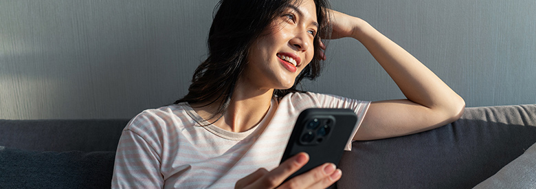 A young woman sits on a sofa, looking up and smiling while holding a smartphone in her hands. 