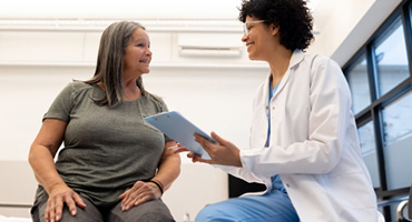  A happy older person with gray hair talks to a smiling medical professional in a white coat while holding a clipboard.