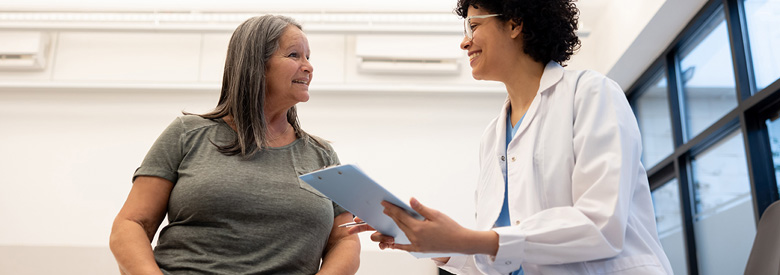  A happy older person with gray hair talks to a smiling medical professional in a white coat while holding a clipboard.