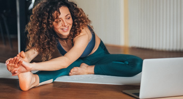 Young adult with curly hair smiles while stretching during an online yoga class on a laptop.