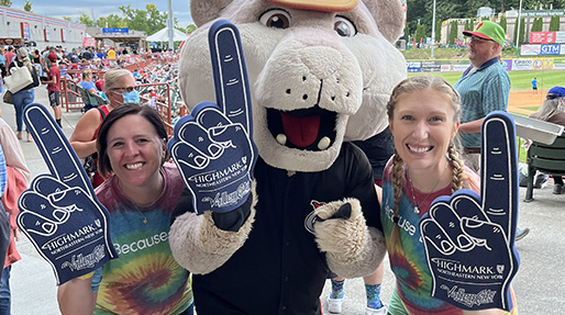 Two adults with bright smiles stand next to a mascot, all holding up big foam fingers with the Highmark Northeastern New York logo at a ball game.