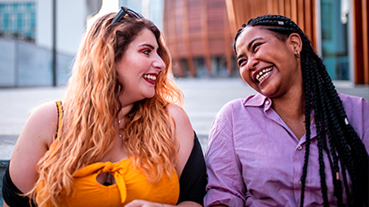 Two women with long hair, one blonde and one dark-braided, sit outdoors on steps, smiling and looking at each other.
