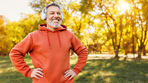 A smiling man with gray hair and a beard, wearing an orange hoodie and white headphones, stands outdoors in a park with golden autumn trees in the background.