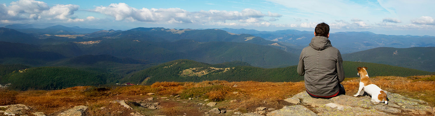 A man in a grey hoodie sits on a rock next to a small brown and white dog, both looking out over a vast mountain landscape under a partly cloudy sky.