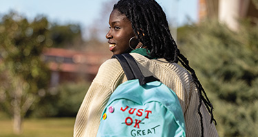 A person wearing a "Just Ok Great" backpack smiles while looking over their shoulder in a bright, outdoor setting.
