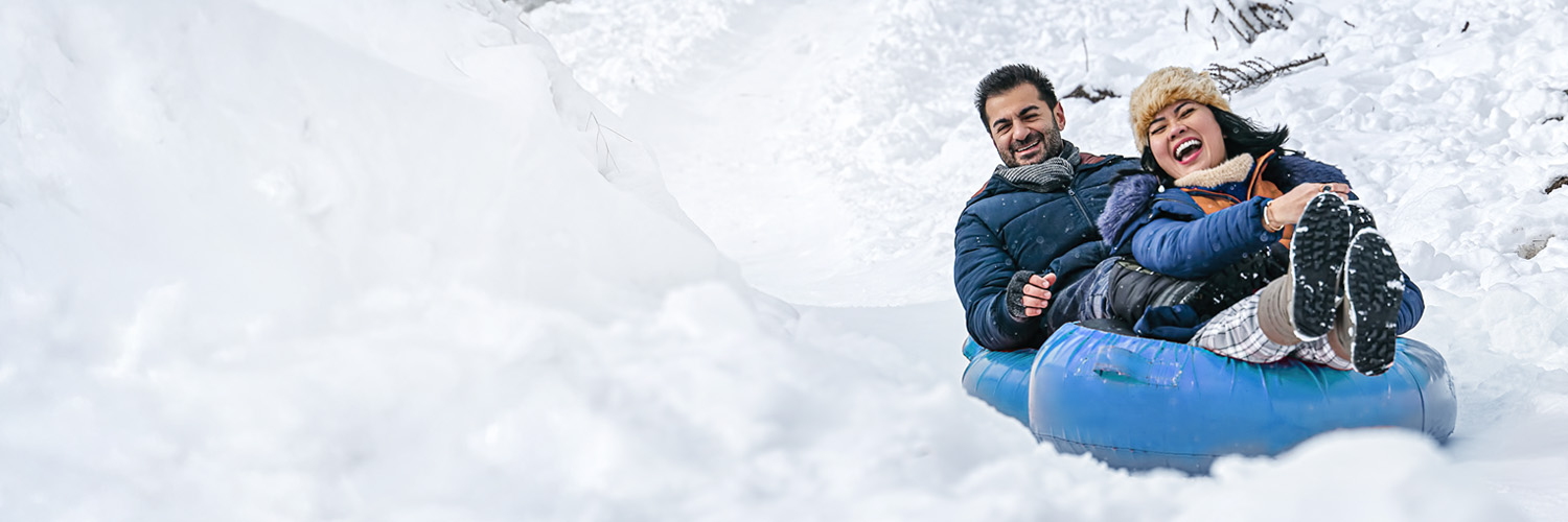 A happy couple bundles up in winter clothes while riding a snow tube down a snowy hill.
