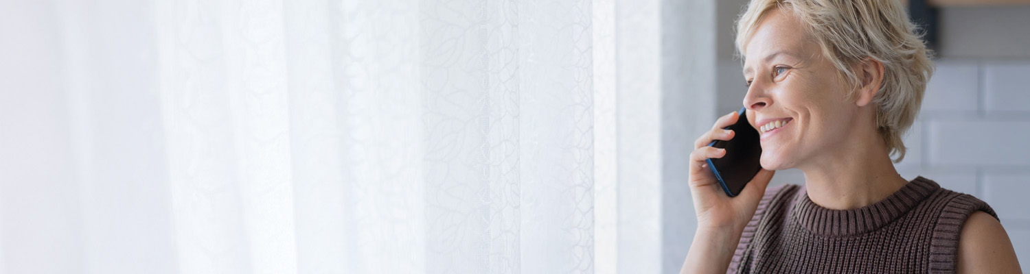 A person with short blonde hair, who looks to be in their 40s, is smiling while talking on a cell phone in front of a window with white, patterned curtains.