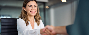 A professional woman with dark hair, wearing a white shirt and blazer, smiling warmly as she shakes hands with another person across a table in an office setting, symbolizing HR collaboration.
