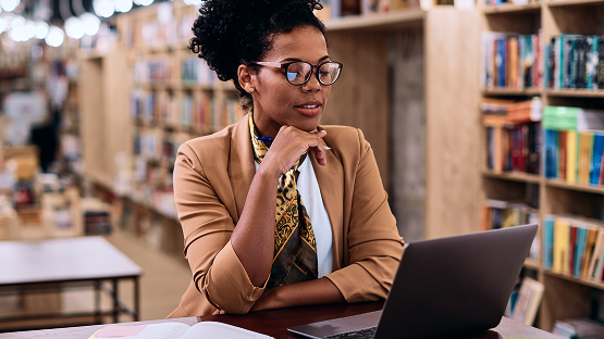 A woman with dark curly hair wearing glasses and a tan blazer, looking thoughtfully at a laptop screen in a library setting.