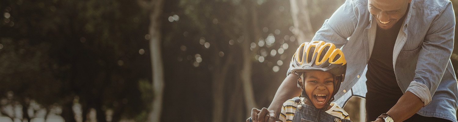 Black man laughing and looking down at young black child with bicycle helmet riding a bicycle to illustrate Highmark CHIP offerings for families. 