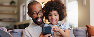 A smiling father holding a smartphone while his young daughter looks at it with interest. This image represents the contact information section for Highmark Healthy Kids.